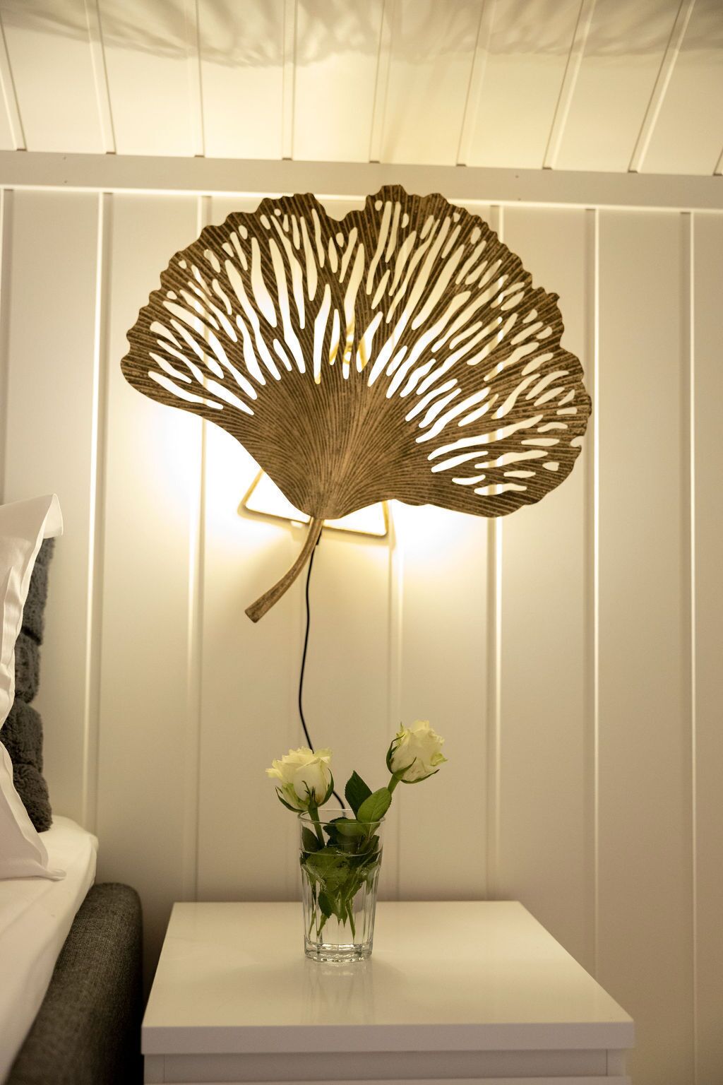 Una habitación con una mesita de noche blanca, un florero de vidrio transparente con flores blancas y un adorno en la pared.