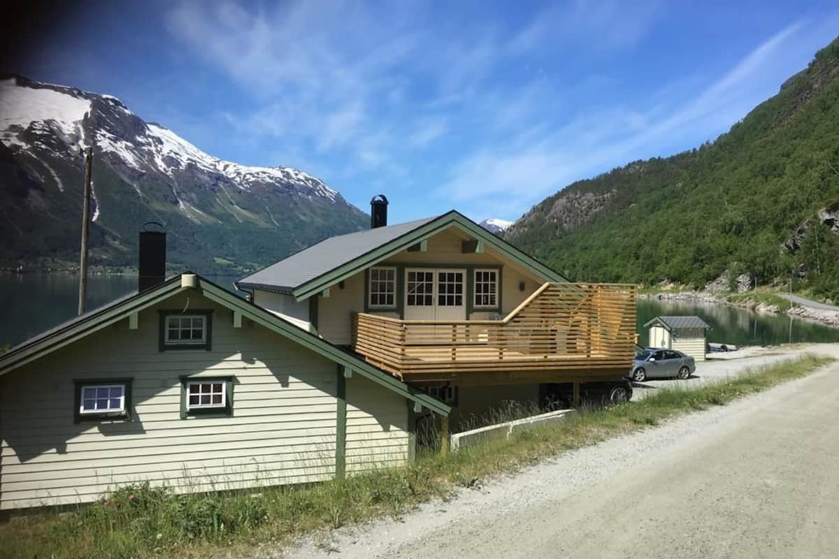 Una casa de dos pisos con un muelle de madera, un coche estacionado al frente y un lago con montañas al fondo.