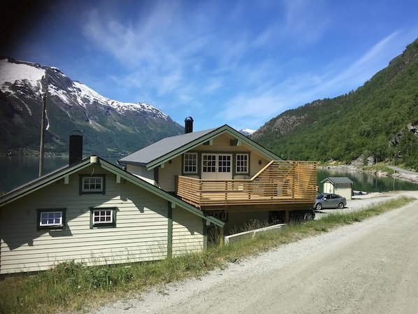 A two-story house with a wooden deck, a car parked in front, and a lake with mountains in the background.
