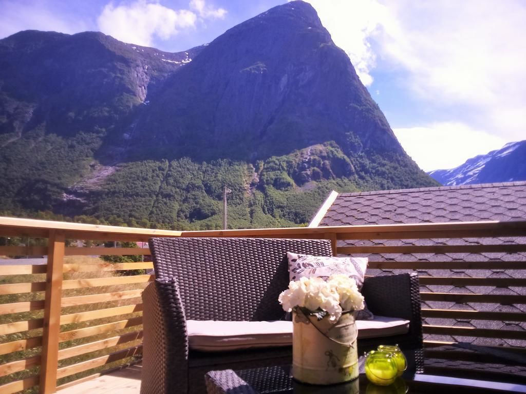 A balcony with wicker furniture, a vase with white flowers, and a glass bottle. A mountain with a peak is visible in the background.
