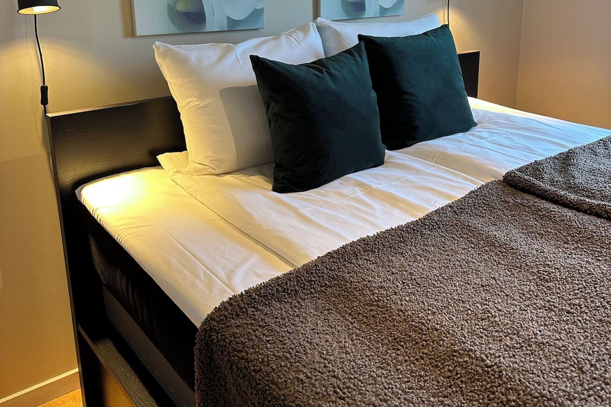 Una cama bien hecha con sábanas blancas y almohadas oscuras, con dos lámparas fijadas a la pared a cada lado.