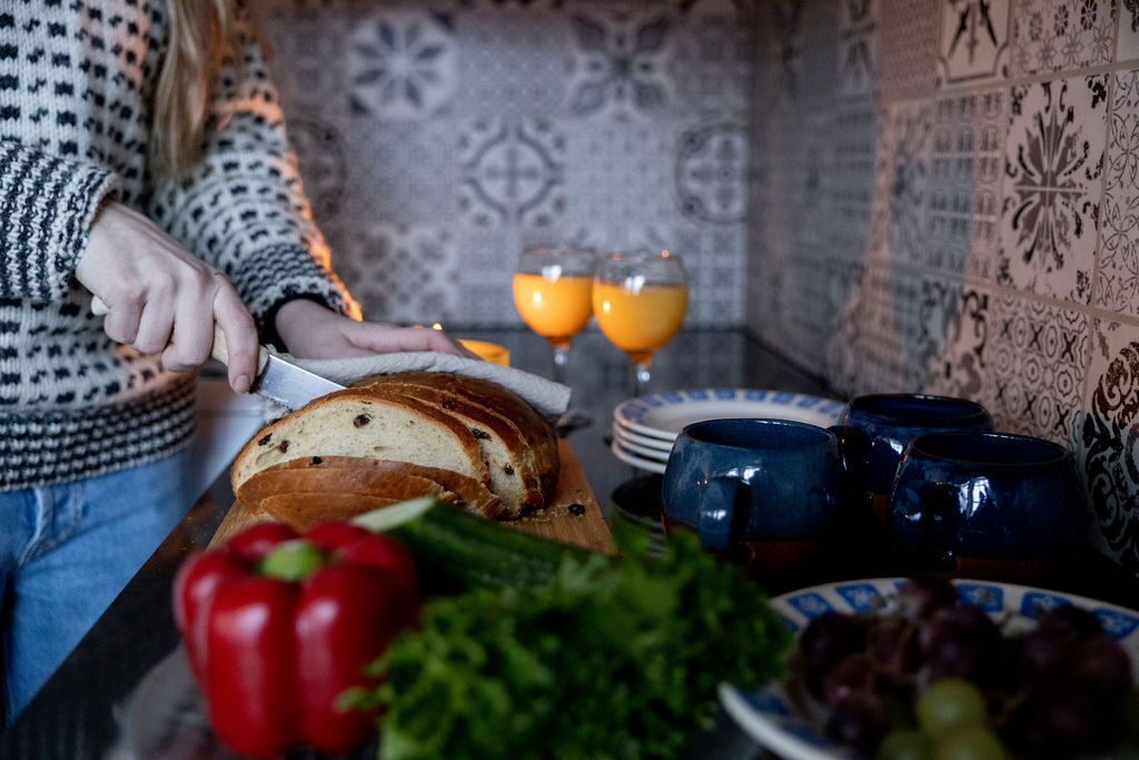 Une personne est en train de trancher du pain sur une planche à découper en bois. On aperçoit sur la table un poivron rouge et des légumes verts, et au fond, deux verres de jus d'orange.