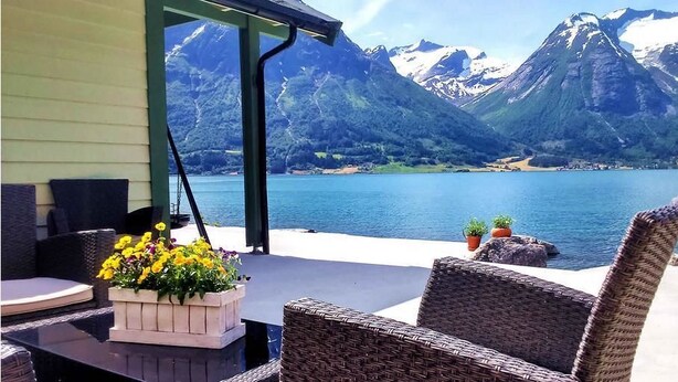 A patio with wicker furniture, a table with a flower arrangement, and a view of a lake with mountains in the background.