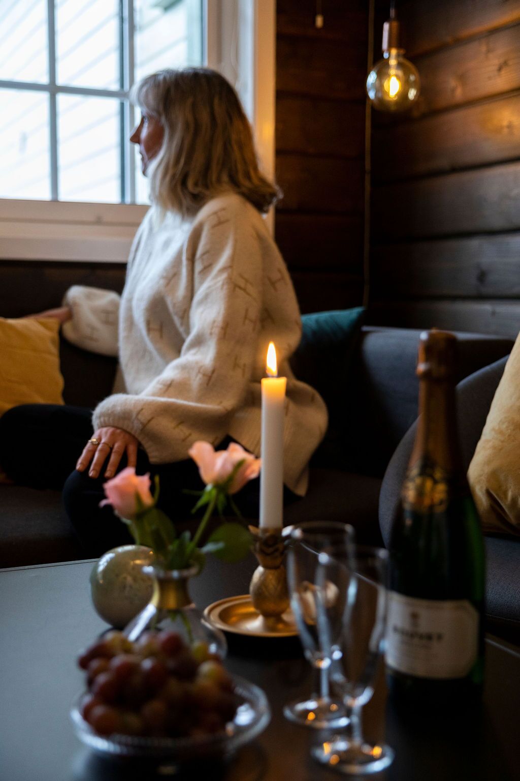 A person sitting on a dark-colored sofa with a candle and a bottle of wine on a table in front of them.