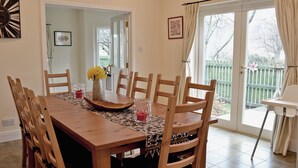 Ein Esszimmer mit einem Holztisch, Stühlen und einer Vase mit Blumen. Ein Fenster mit Vorhängen bietet einen Blick ins Freie.