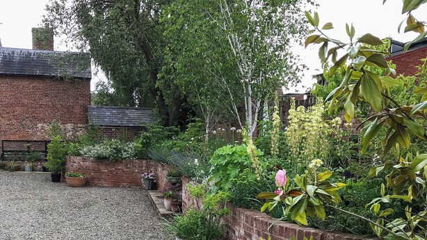 Ein Garten mit Kiesweg, Backsteinmauern und verschiedenen Kübelpflanzen.
