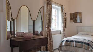 Ein Schlafzimmer mit einem hölzernen Schminktisch, zwei Betten mit gemusterten Bettbezügen, einem großen Spiegel und einem Fenster mit blumigen Vorhängen.