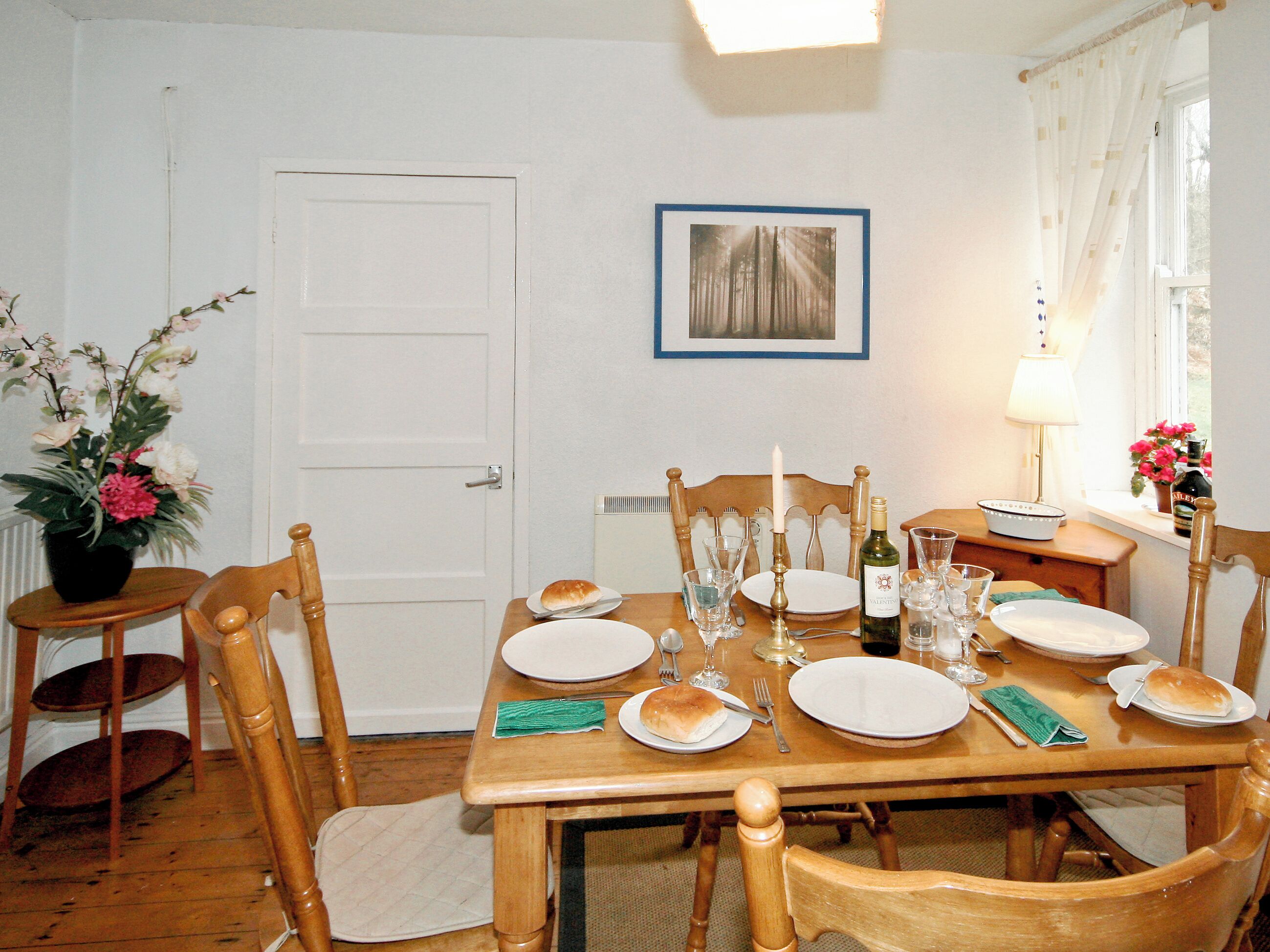 Un comedor con una mesa de madera dispuesta para cuatro personas, sillas, un florero con flores, un cuadro en la pared y una ventana con cortinas.
