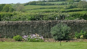 A stone wall with a flower bed and a grassy field in the foreground. - 2 bedroom accommodation in Shipton Gorge (Shipton Gorge)