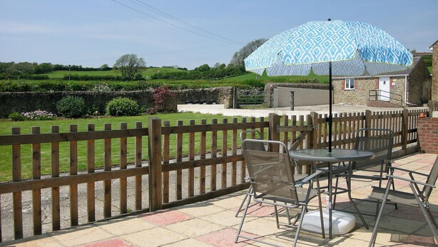 An outdoor patio with a table and chairs, a patterned umbrella, and a view of a rural landscape with a stone building in the background. - 2 bedroom accommodation in Shipton Gorge (Shipton Gorge)