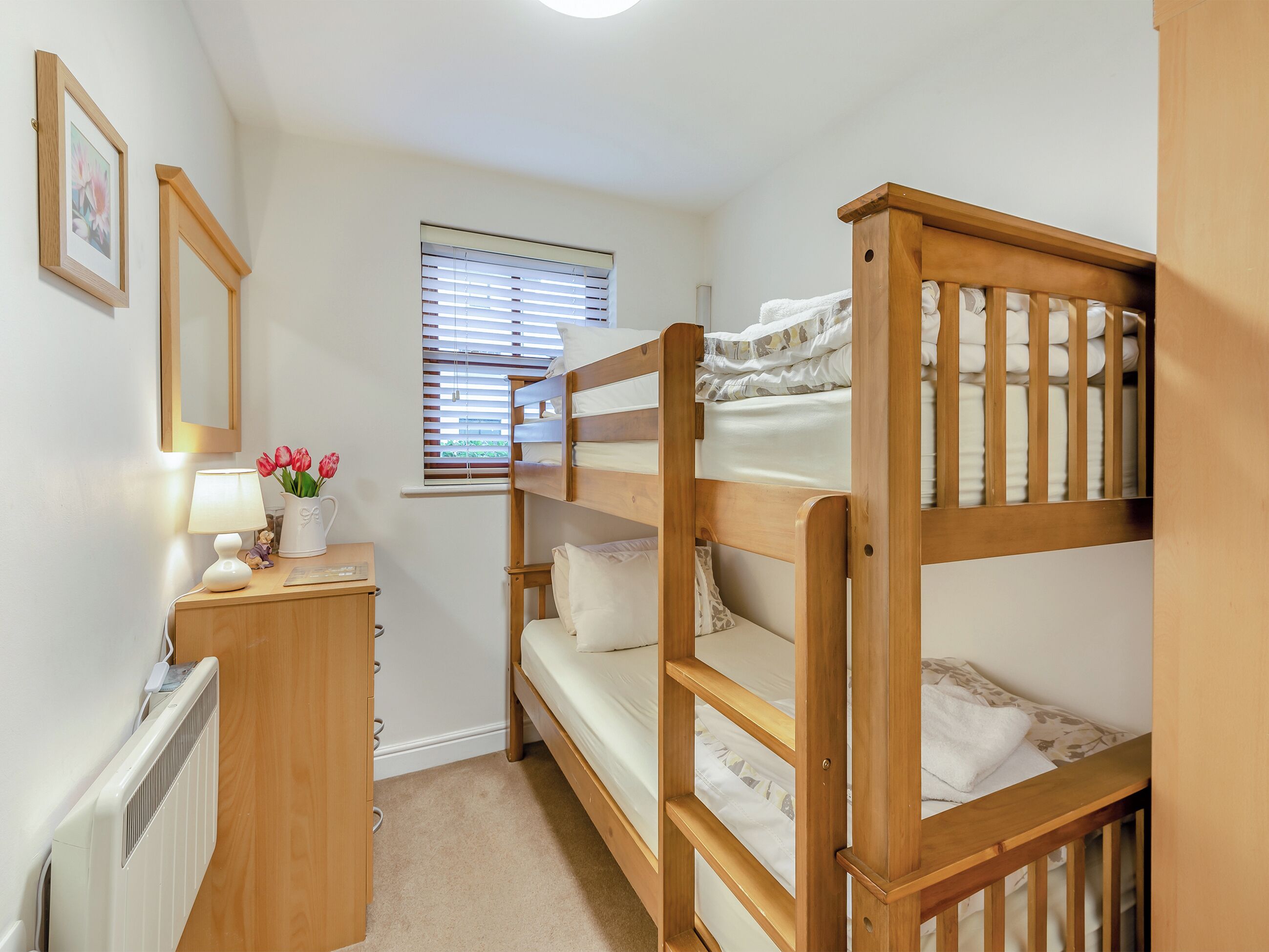 A bunk bed room with a desk, a radiator, a window with blinds, and a framed picture on the wall.
