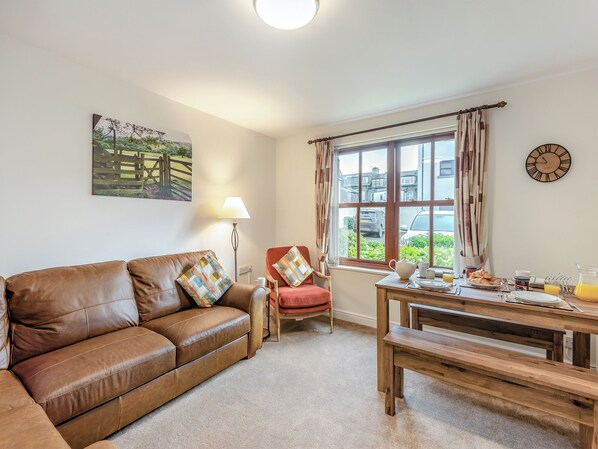 A cozy living room with a brown leather sofa, a red armchair, a wooden dining table with breakfast items, a large window with curtains, and a wall clock. - 2 bedroom accommodation in Keswick (Keswick)