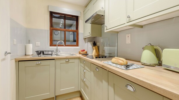 A kitchen with a sink, countertop, and cabinets. There is a window with a clock, a toaster, and a bread loaf on the counter.
