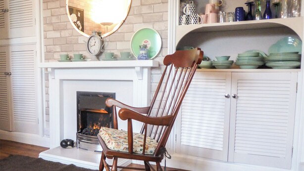 A rocking chair in front of a fireplace with a brick wall and white shutters.