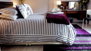 A neatly made bed with striped bedding and multiple pillows, a desk with a lamp, a chair, and a mirror.