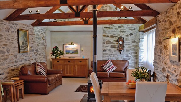 A cozy living room with stone walls, wooden beams, leather sofas, a wooden dining table, and a fireplace. - Penno Cottage - 24440 (Bodmin)