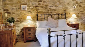 A stone-walled bedroom with a bed, two bedside tables with lamps, a wooden nightstand, and a small table with a plant. - Penno Cottage - 24440 (Bodmin)