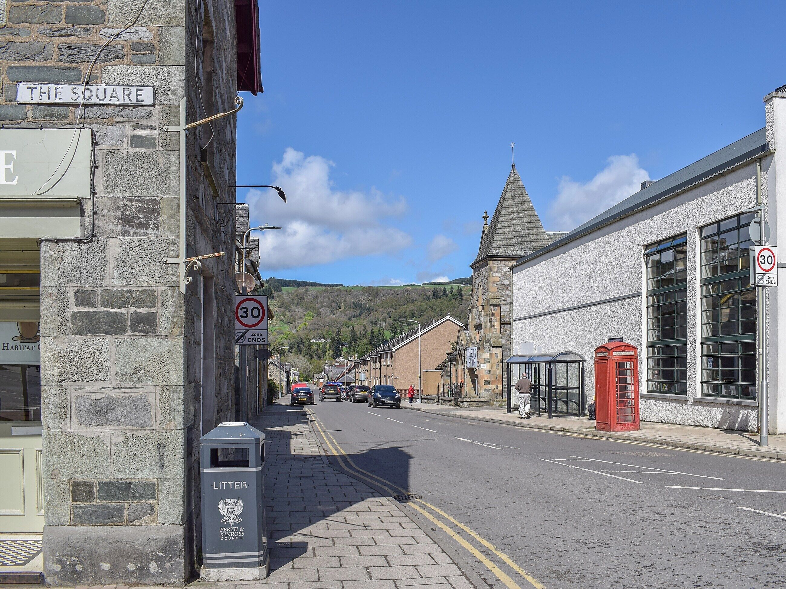 A street view with a stone building labeled "THE SQUARE," a red telephone booth, and a church with a pointed roof.
