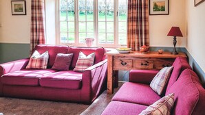 A living room with a red sofa set, a wooden side table, and a window with curtains. - Moorhouse Farm Cottage (Hovingham, near York)
