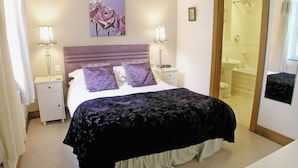 A bedroom with a bed, two bedside tables, a nightstand, a bathroom with a shower, and a door leading to another room.