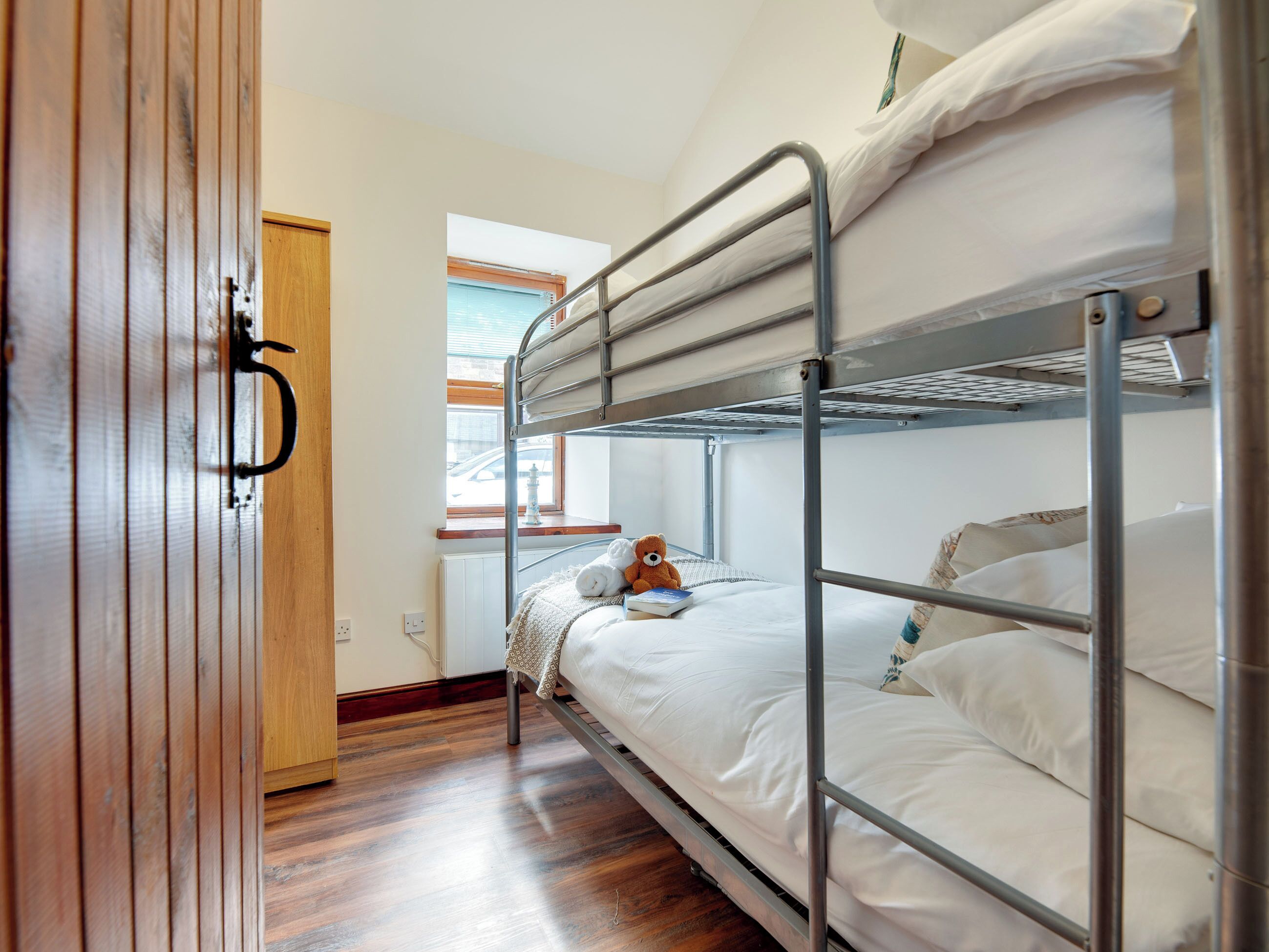 A bunk bed room with a wooden door, a small window, and a teddy bear on the bed.