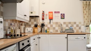 Une cuisine avec des meubles blancs, un grille-pain, une machine à café et un lave-vaisselle Bosch. On y trouve également des ustensiles de cuisine, une couverture anti-feu et un extincteur fixés au mur.