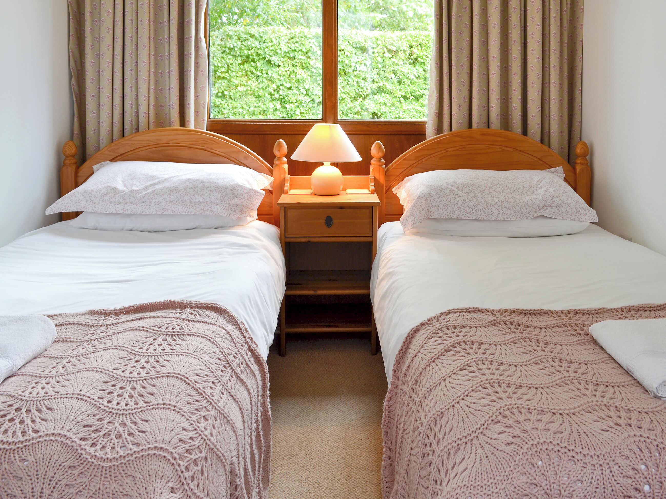 A room with two single beds, a nightstand with a lamp, and a window with curtains.