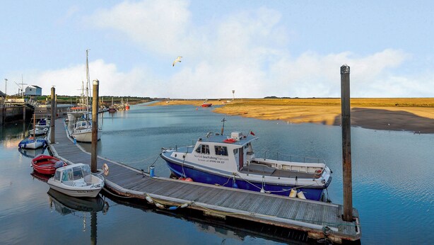 Een jachthaven met afgemeerde boten, een houten steiger en een zandstrand op de achtergrond.