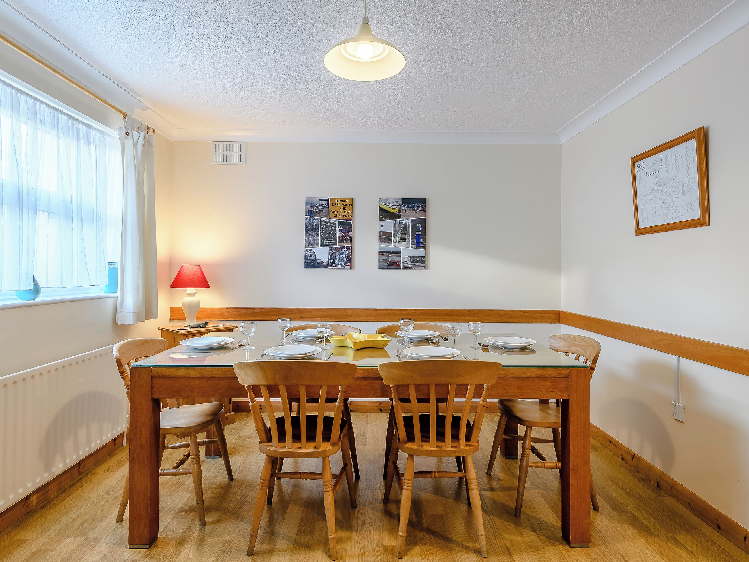A dining area with a table set for four, wooden chairs, a hanging light fixture, a window with curtains, and a wall-mounted picture.