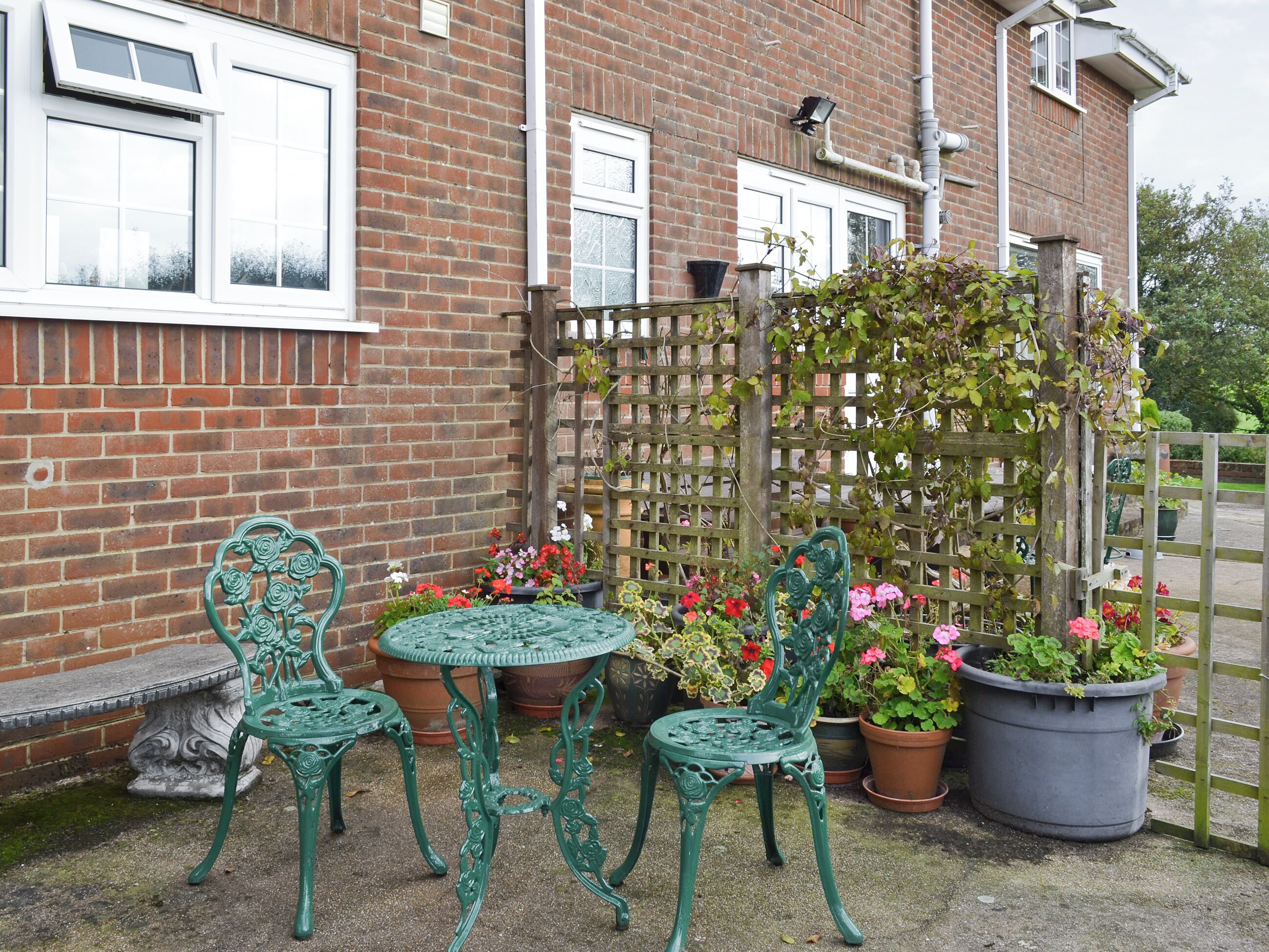 A brick building with a garden area featuring green metal chairs, a small round table, and potted plants with flowers.