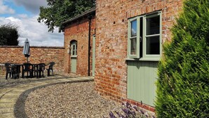 A brick building with a green door, a window with a white frame, and a patio area with a table and chairs. - The Bran House- 23526 (Hareby, near Horncastle)
