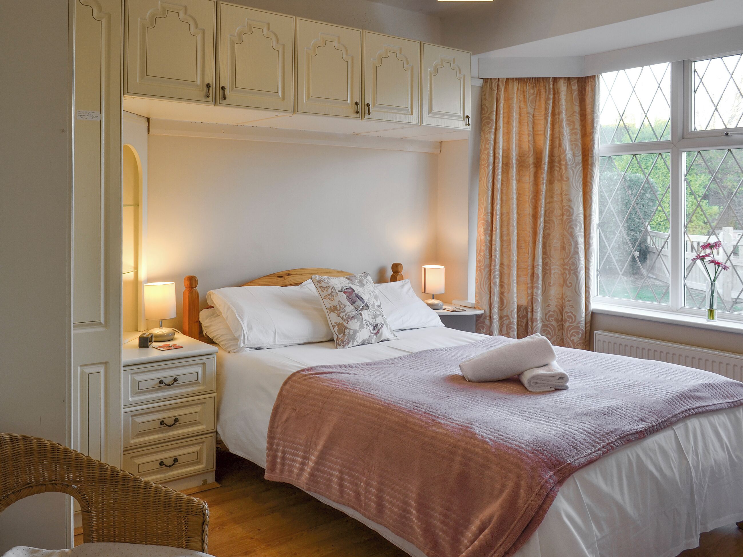 A bedroom with a large bed, two bedside tables with lamps, a chest of drawers, a wicker chair, and a window with curtains.