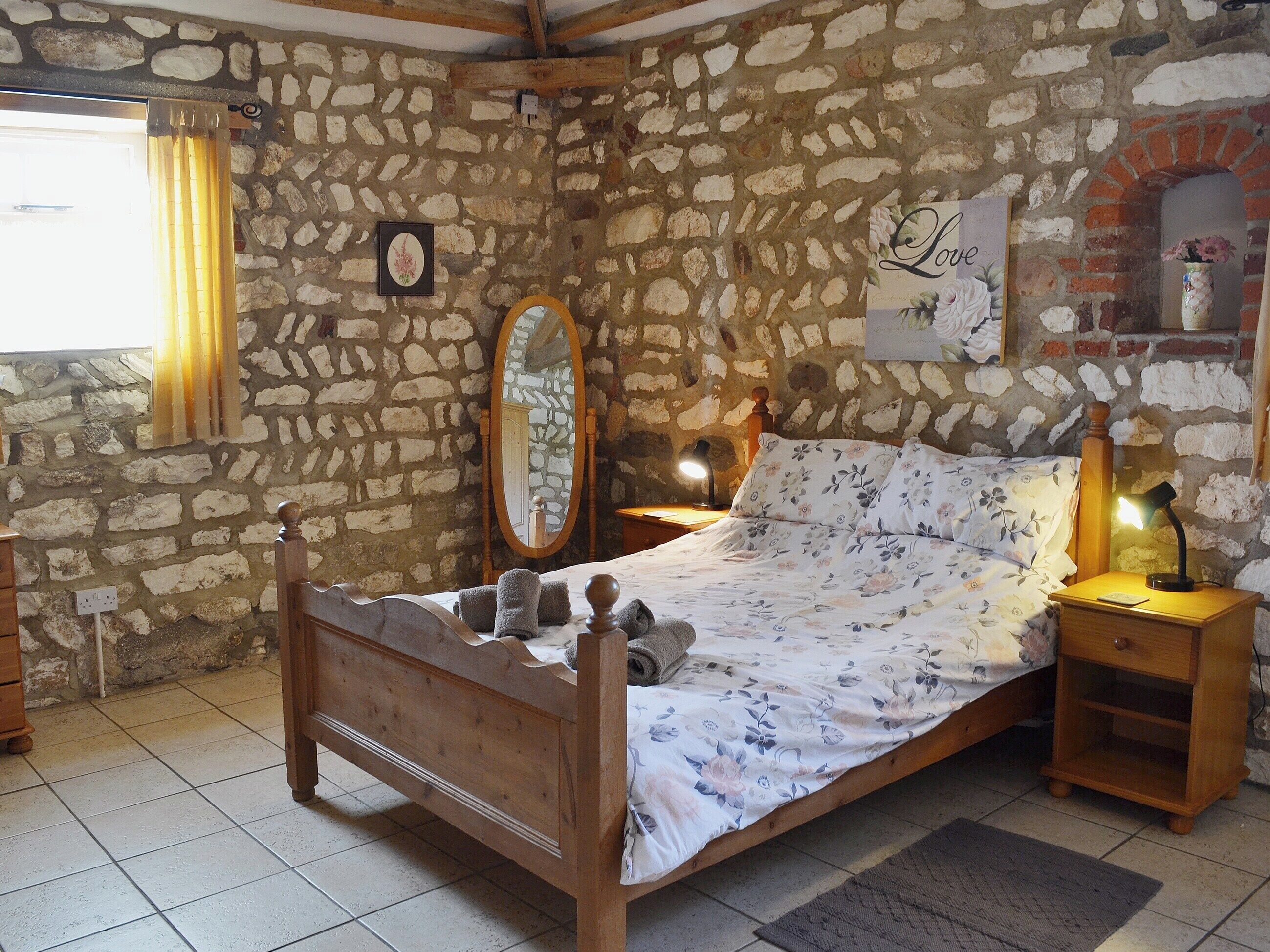 Ein Schlafzimmer mit Steinwänden, einem hölzernen Bett, einem Nachttisch mit Lampe und einem Spiegel an der Wand.