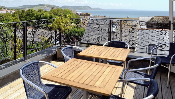 Une terrasse dotée de deux tables en bois et de quatre chaises en osier, offrant une vue panoramique sur la côte, avec ses collines et la mer.