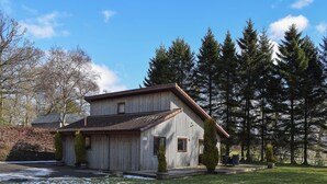 Ein rustikales Holzgebäude mit geneigtem Dach, umgeben von einer grasbewachsenen Fläche mit Schneeflecken, und dichten immergrünen Bäumen im Hintergrund.