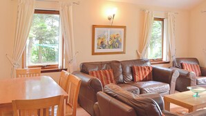 Ein Wohnzimmer mit braunem Ledersofa, einem Holzesstisch und Stühlen. Zwei Fenster mit Vorhängen, ein gerahmtes Bild an der Wand und eine Leuchte sind vorhanden.