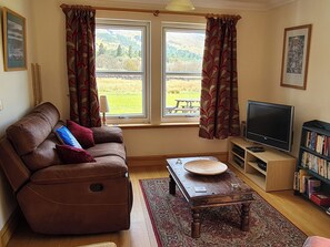 A living room with a brown leather sofa, a wooden coffee table, a television on a stand, a bookshelf, and a window with curtains. - 3 bedroom accommodation in Ardgour, near Fort William (Ardgour, near Fort William)