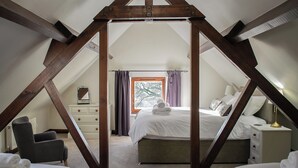 A bedroom with a sloped ceiling, wooden beams, a bed, a chair, a dresser, and a window with curtains. - Characterful, rural barn conversion. (Heathcote, near Hartington)