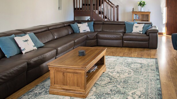 A living room with a brown leather sectional sofa, a wooden coffee table, a patterned rug, and a staircase in the background. - Characterful, rural barn conversion. (Heathcote, near Hartington)