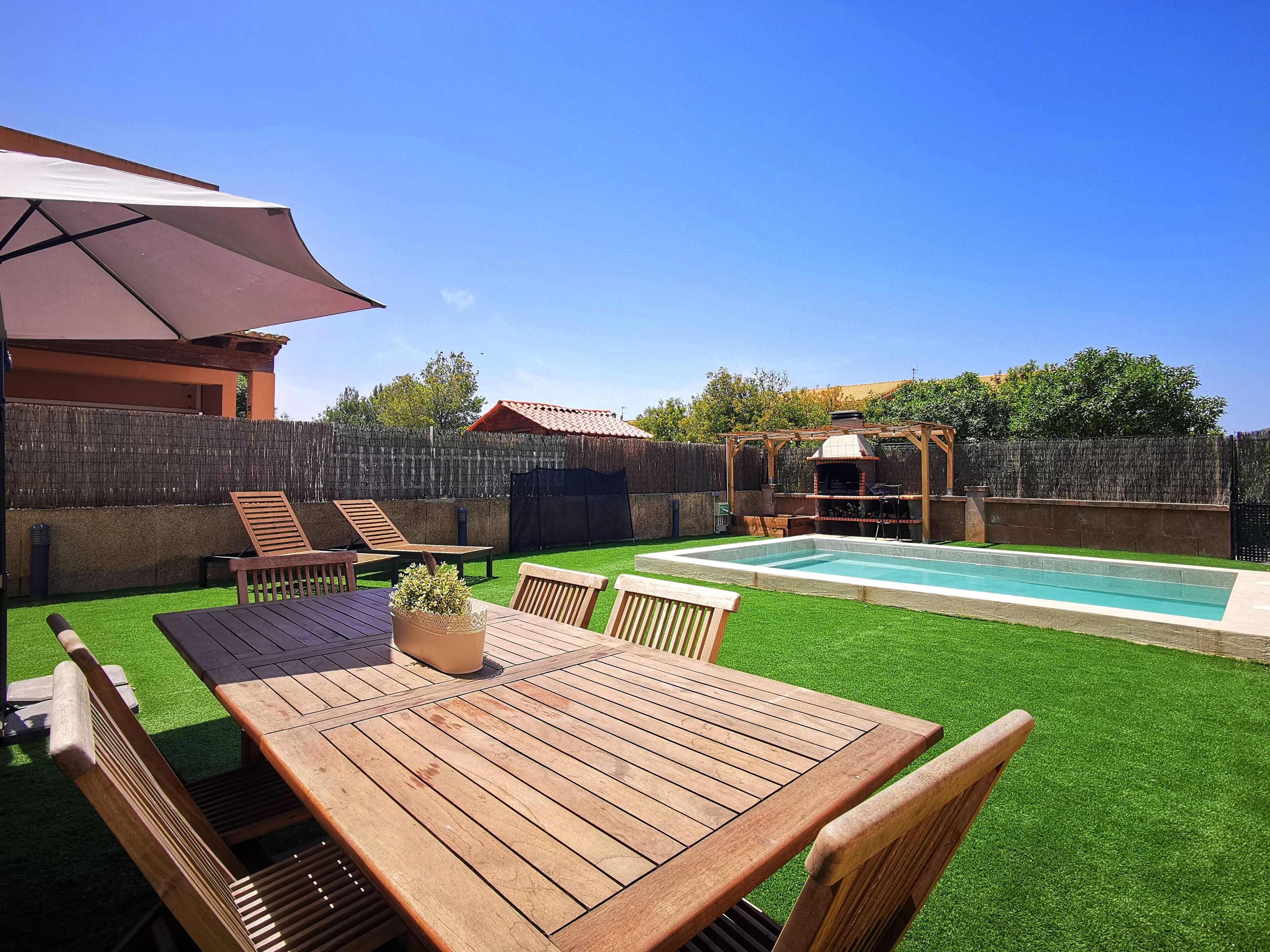 Un patio trasero con mesa y sillas de madera, una parrilla de barbacoa y una piscina.