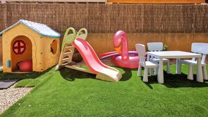 Un'area giochi per bambini con uno scivolo, un gonfiabile a forma di fenicottero, una casetta e tavoli e sedie bianche.