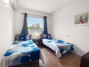 A bedroom with two single beds, a nightstand with a lamp, a window with curtains, and a painting on the wall.