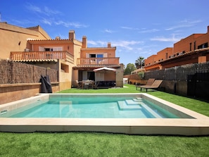 Una piscina con zona de patio, área de estar cubierta y vista a un edificio de varios pisos con balcones.