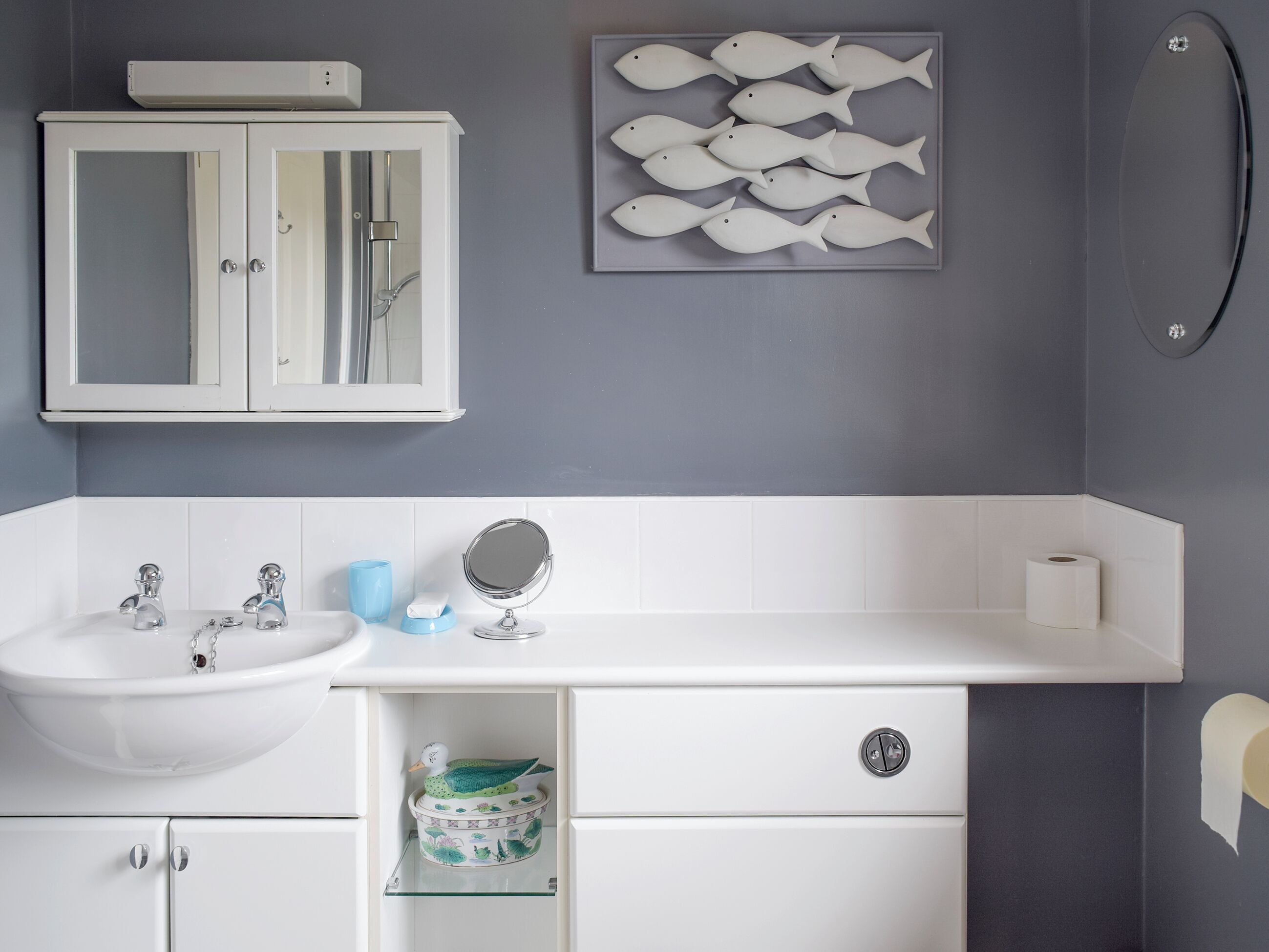 A bathroom with a white sink, two faucets, a mirror, a toilet paper holder, and a cabinet with a glass shelf.