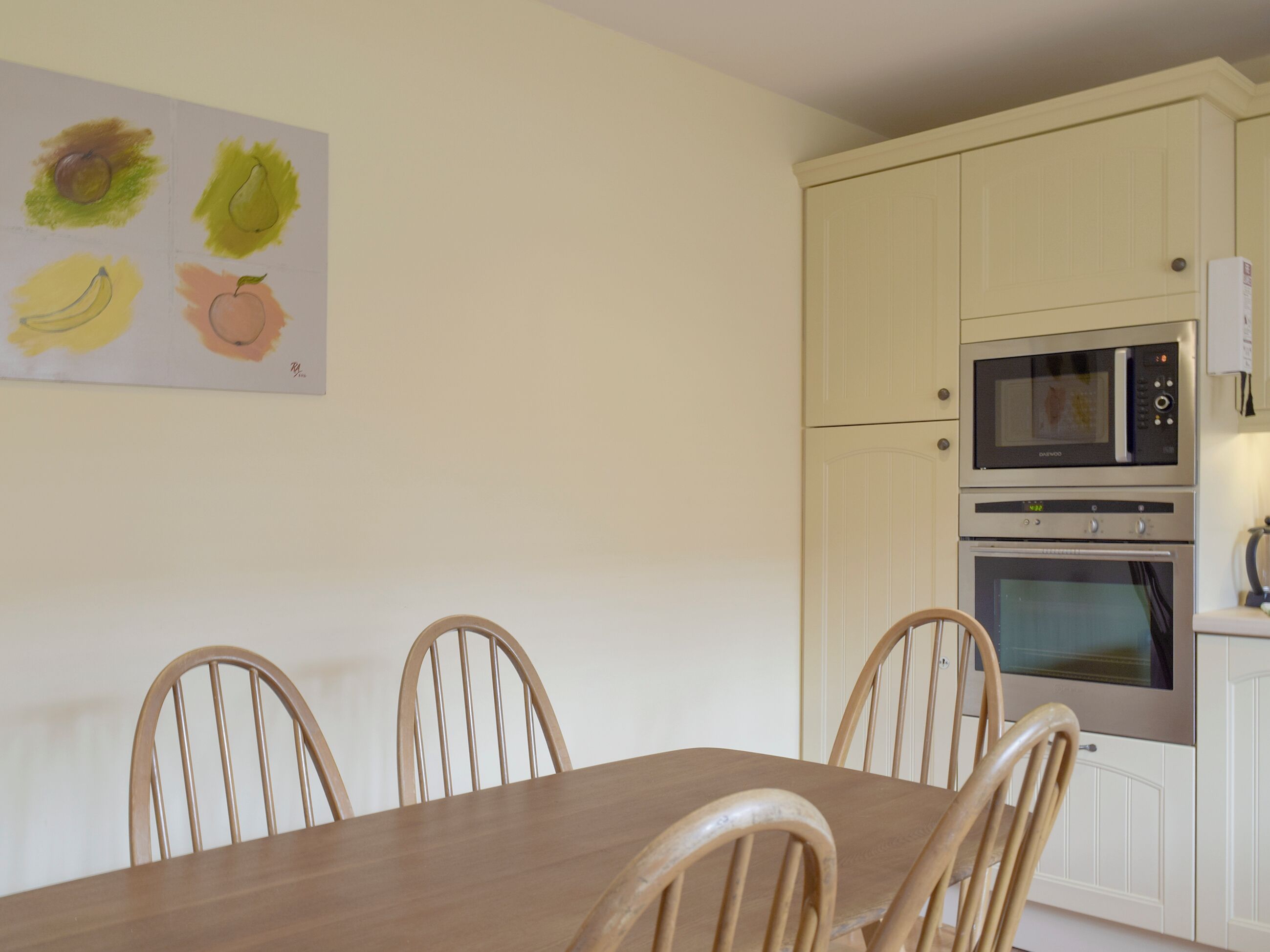 Un coin repas avec une table en bois et quatre chaises, une cuisine avec des meubles blancs et deux fours, et un tableau représentant des fruits accroché au mur.