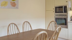 Un comedor con una mesa de madera y cuatro sillas, una cocina con armarios blancos y dos hornos, y un cuadro de fruta colgado en la pared.
