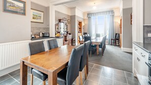 A dining area with a wooden table and chairs, a kitchen with modern appliances, and a large window with curtains. - 7 bedroom accommodation in Portpatrick (Portpatrick)
