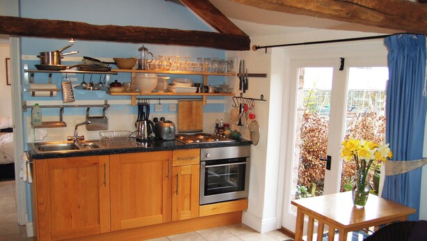 A kitchen with wooden cabinets, a stainless steel sink, and a built-in oven. There is a dining area with a table and chairs, and a window with blue curtains. - A delightful little barn conversion situated in a tranquil area of Cumbria. (Ellonby)