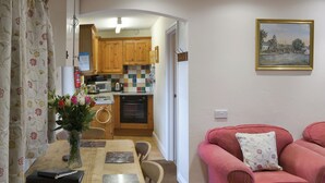 A dining area with a wooden table, chairs, a vase with flowers, and a red sofa with a floral pillow. - A pretty 2 bedroom barn conversion set within an equestrian centre. (Polson, nr. Launceston)