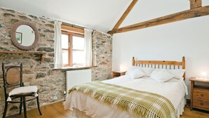 Quarto com parede de pedra, vigas de madeira, cama com roupa de cama branca, uma cadeira, mesa de cabeceira e um espelho.